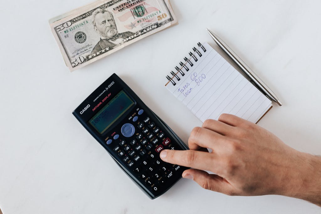 A hand using a calculator next to a notepad and cash, representing financial planning.