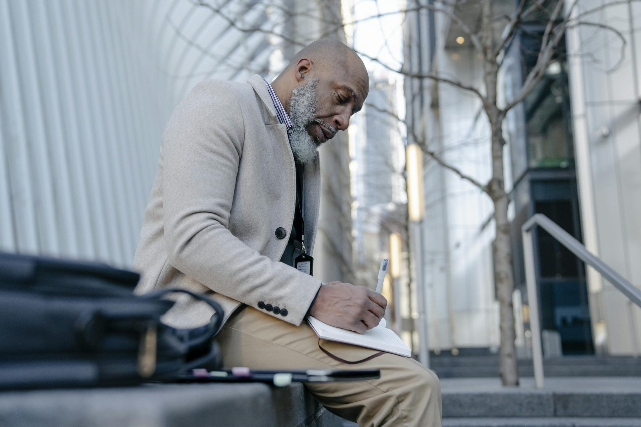 An elderly man in a suit sits outdoors writing in a notebook, capturing a moment of focus in an urban setting.