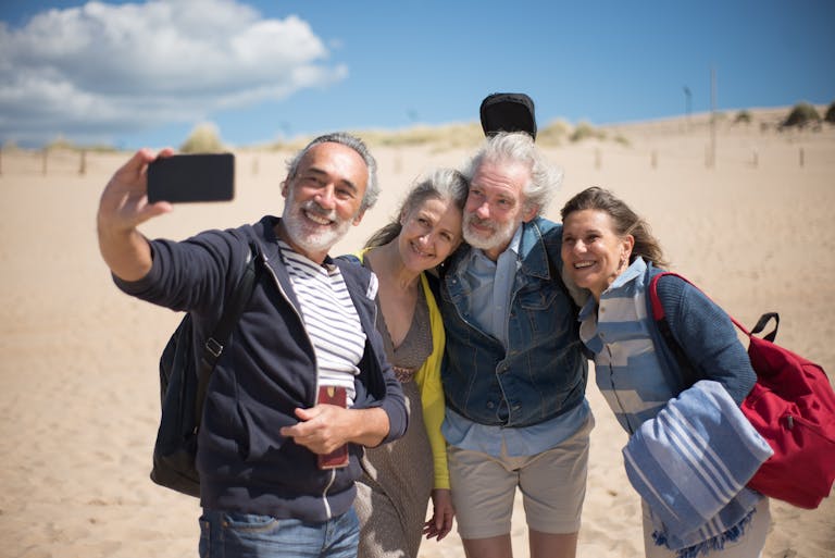 A joyful group of senior friends taking a selfie at a sandy beach after using travel savings tips from Thrifty Boomers