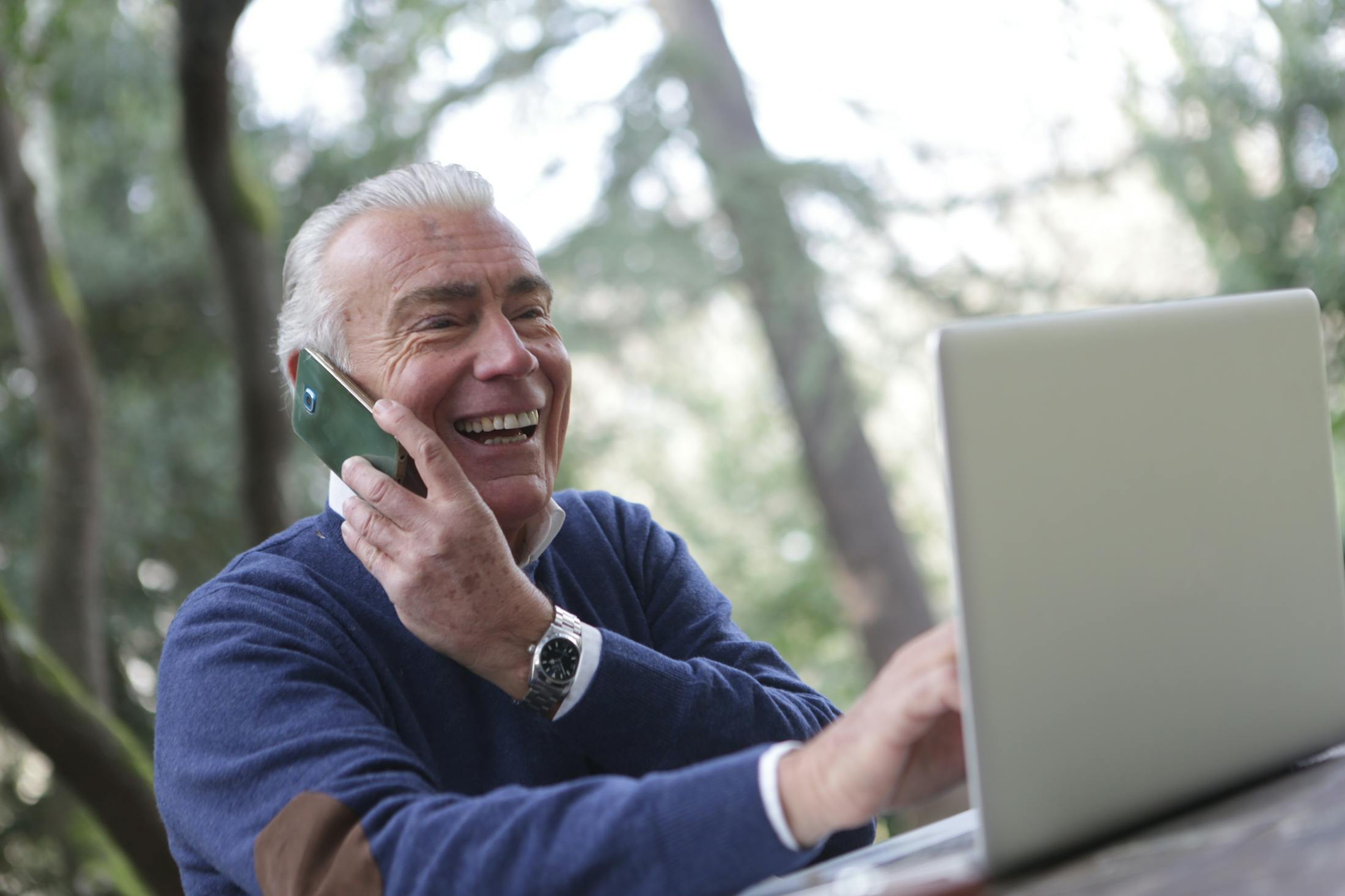 Happy retired boomer man working on side hustle from laptop making extra money after 60 while talking on the phone