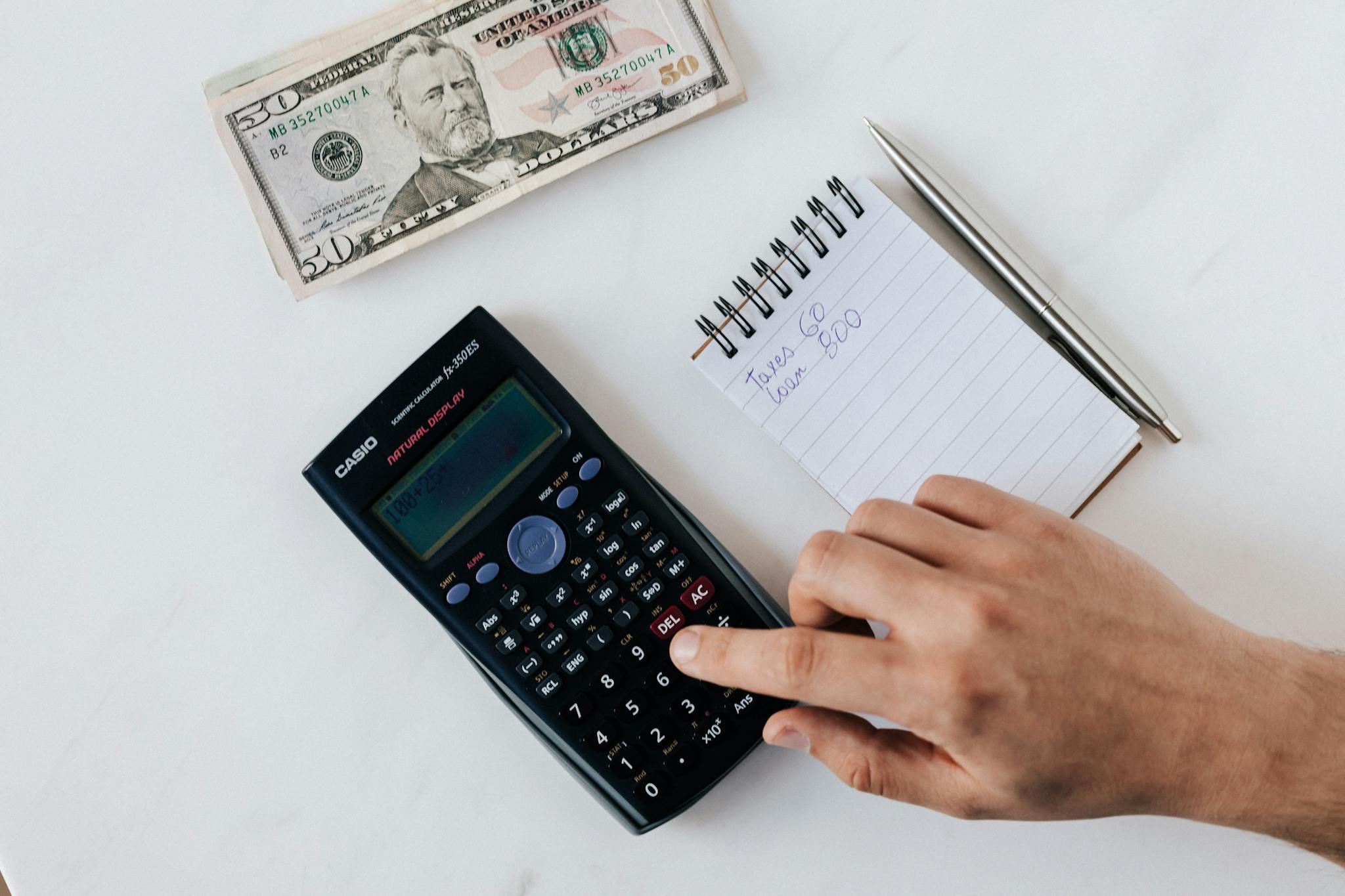 A hand using a calculator next to a notepad and cash, representing financial planning.